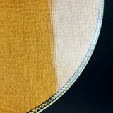 Close-up of a guitar body showing the natural wood grain top, the white inner rim, and a decorative gold and black inlay border against a dark background.