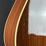 Close-up of a wooden acoustic guitar body showing a rich reddish-brown wood grain finish, a gold-toned binding edge, and a glossy reflective shell on a dark background.