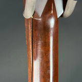 Close-up of a polished dark brown wooden gunstock with a silver metal butt cap and a vertical silver barrel shroud, set against a dark gray background.