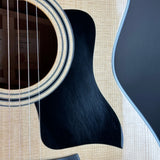 Close-up detail of an acoustic guitar body showing the natural wood grain top, sound hole with rosette, black pickguard, and part of the dark side panel.