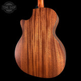 Close-up of the back of a wooden acoustic guitar body with a rich warm brown finish and prominent grain pattern, shown against a solid black background with a Tone Shop watermark in the top left.
