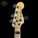Close-up of a Fender Jazz Bass headstock in natural wood with four black tuning pegs and a black logo, shown against a solid black background. A Tone Shop watermark is visible in the top left corner.