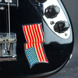 A close-up of a glossy black electric guitar body featuring a waving American flag enamel pin on the side, with black control knobs and a chrome bridge visible.
