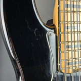 Close-up of a black electric bass guitar body with visible scratches on the glossy finish, showing part of the gold-toned fretboard and strings.