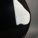 Close-up of a glossy black curved surface with a bright white reflection and a small dark defect or hole near the center, against a gray background.