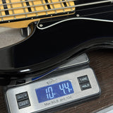 A close-up of a black electric bass guitar resting on a digital scale, showing the scale's blue display screen and control buttons.