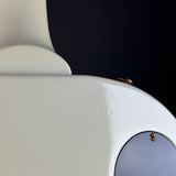Close-up detail of a white electric guitar body showing a gold-toned screw, a circular silver plate, and part of the neck against a dark background.