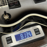 A close-up of a guitar body resting on a digital scale displaying 8.134 oz.