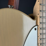 Close-up detail of an electric guitar body in a warm beige/tan finish, showing part of the neck, strings, and a light blue pickguard.