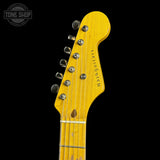 Close-up of the headstock of a Fender Stratocaster electric guitar with a natural yellow maple wood finish, chrome tuners, and six strings, shown against a solid black background.