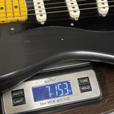 A close-up of a black electric guitar body resting on a digital scale displaying 7.153 oz, with the guitar's white pickups and yellow headstock partially visible.