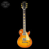 A Gibson Les Paul Custom electric guitar in a sunburst finish, shown from the front against a solid black background. The guitar features a warm orange-to-yellow sunburst body, a cream pickguard, gold hardware, and a dark fretboard with gold block indicators.