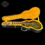 An electric guitar with a metallic olive green body and black neck, displayed inside an open black hard case with a bright yellow-orange velvet interior, set against a solid black background.