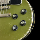 Close-up of an electric guitar body in distressed olive green, showing four black control knobs with silver centers, a black pickguard, and a gold-toned edge trim against a black background.