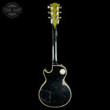A vintage black electric guitar shown from the back, featuring a glossy black finish with gold trim and a worn, scuffed body. The headstock is light green with gold tuning pegs.