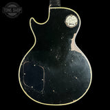 The back of a distressed black Gibson Les Paul Custom electric guitar, showing heavy wear, scuffs, and a faded gold outline around the body contour.