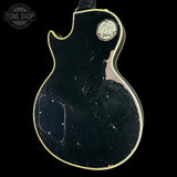 A vintage Gibson electric guitar body, likely a Les Paul Custom style, shown from the back against a solid black background. The body features a distressed black finish with heavy wear, scuffs, and rust, highlighted by a gold-toned binding.