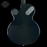 The back of a glossy black electric guitar, showing the cutaway body shape, two circular pickups, and a clear plastic cover over one pickup, set against a solid black background.
