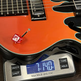Close-up of an orange electric guitar body resting on a digital scale, with a small red die placed on the guitar surface. The scale reads 7.