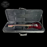 A red electric guitar resting inside an open black hard case with a gray velvet interior, photographed against a solid black background. A Tone Shop watermark is visible in the top-left corner.