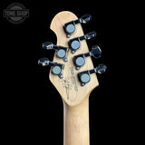 Close-up of the headstock of a natural wood electric guitar against a black background, featuring six black tuning pegs with silver accents and a Tone Shop logo watermark in the top left corner.
