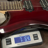 A close-up of a glossy dark red electric guitar resting on a digital scale, showing the scale display reading 7.64 oz.