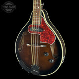 A close-up studio shot of a mandolin against a solid black background. The mandolin features a dark brown sunburst finish, a vibrant red marbled pickguard, cream-colored binding, and a black bridge.