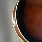 Close-up detail of an acoustic guitar's body corner, showing the dark brown wood finish, a cream-colored binding edge, and a silver metal plate near the sound hole on a neutral gray background.