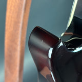 Close-up detail shot of a glossy dark reddish-brown electric guitar body corner, showing the high-gloss finish and part of a wooden neck or headstock in the background.