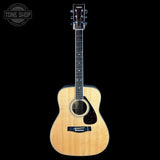 A natural wood acoustic guitar with a dark headstock and black pickguard, shown vertically against a solid black background. A Tone Shop watermark is visible in the top-left corner.