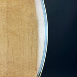 Close-up detail of a guitar body showing the natural wood grain on the back and the edge of the soundboard with blue and white binding against a dark background.
