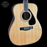 A close-up of an acoustic guitar body with a natural flamed wood top, black pickguard, and dark sides, shown against a black background with a Tone Shop watermark in the upper left.