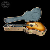 An acoustic guitar with a natural sunburst finish resting inside an open hard case with blue felt lining, shown against a solid black background. A Tone Shop watermark is visible in the top-left corner.