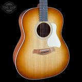 A close-up studio shot of an acoustic guitar body against a black background, showing a natural sunburst finish that fades from bright amber-orange at the edges to a darker brown center, with a white pickguard and a visible Tone Shop logo watermark in the top-left corner.