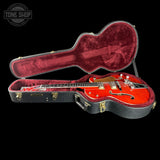 A red electric guitar with gold hardware displayed inside an open black hard case with a burgundy velvet interior, set against a solid black background. A Tone Shop logo is visible in the top-left corner.