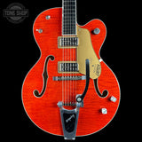 An electric guitar with a vibrant orange-red maple top, gold pickguard, and silver hardware, shown against a solid black background. A Tone Shop watermark is visible in the top-left corner.