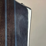 Close-up of a guitar or stringed instrument body corner showing a dark wood grain section, a lighter wood grain section, and a glossy black inlay with visible scratches, against a plain beige background.
