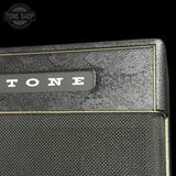 Close-up of a Tone brand guitar amplifier cabinet corner, showing a textured gray fabric grille and a gray nameplate with white TONE lettering, set against a black background.