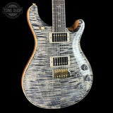 A close-up studio shot of an electric guitar body against a black background, showing a flamed maple top with a blue-gray and white swirling pattern, gold hardware, and a dark fretboard. A Tone Shop watermark is visible in the upper left corner.