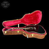 An open brown acoustic guitar case with bright red velvet lining, holding a black acoustic guitar with gold hardware, shown on a black background. A Tone Shop logo is visible in the top-left corner.