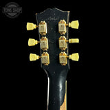Close-up of the headstock of an electric guitar against a black background, showing a black neck with gold tuning pegs and a partially visible natural wood fingerboard at the bottom. A Tone Shop watermark is visible in the top left corner.