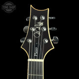 Close-up of a guitar headstock with six tuning pegs, featuring a black finish, gold outline, white script branding, and a white logo on a black background.