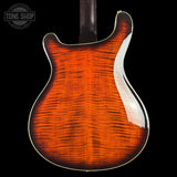 Close-up of the back of a double-cutaway electric guitar showing a vibrant flamed maple top with a classic sunburst finish that transitions from bright orange in the center to dark brown and black at the edges, with a white binding edge. A Tone Shop watermark is visible in the top left corner.