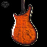Close-up of the back of an electric guitar body showing a vibrant flamed maple wood finish with a dark brown to amber-orange gradient and cream binding, set against a solid black background.