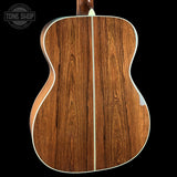 The back of an acoustic guitar featuring a rich, warm brown wood grain finish with a central cream-colored binding and soundhole ring, set against a solid black background.