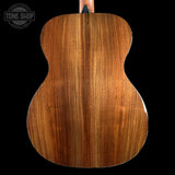 Close-up of the back of an acoustic guitar showing a rich, warm brown wood grain finish against a solid black background, with a Tone Shop logo in the top left corner.