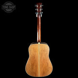 The back view of an acoustic guitar with a natural wood finish, featuring a dark reddish-brown neck and a light-colored center stripe, set against a solid black background with a Tone Shop watermark in the top left corner.