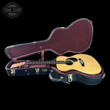 An acoustic guitar with a natural wood top and dark neck, displayed inside an open black hard case with burgundy lining, set against a solid black background. A Tone Shop logo is visible in the top-left corner.