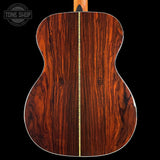 The back of an acoustic guitar made of rich, dark reddish-brown wood with a central gold-toned inlay, shown against a solid black background. A Tone Shop watermark is visible in the top-left corner.