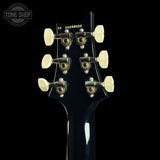 Close-up rear view of a guitar headstock in glossy black, showing six cream-colored tuning keys with gold-toned screws and gold model numbers printed on the back.